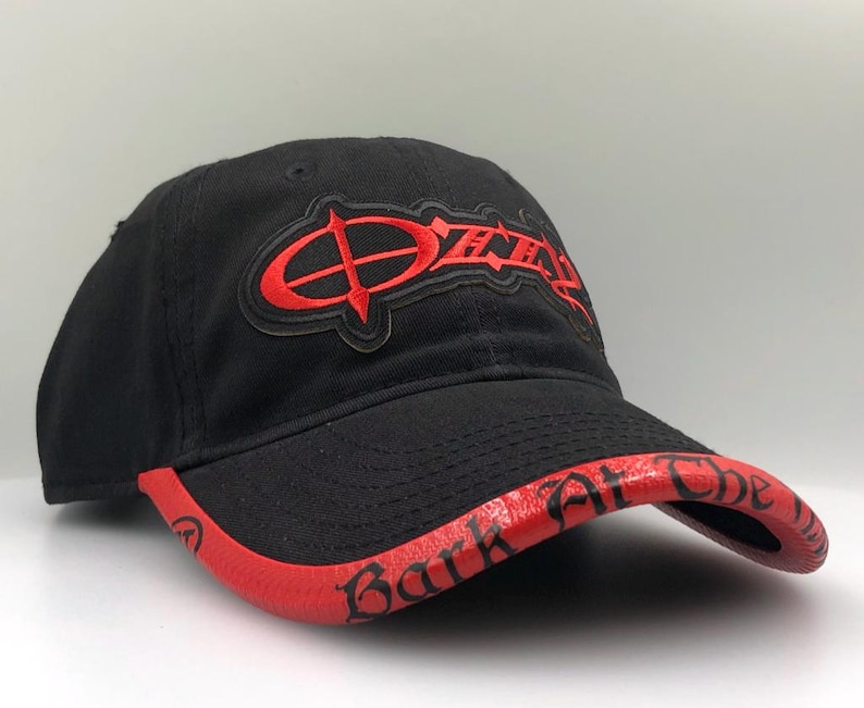 May include: Black baseball cap with a red brim and embroidered red text. The text on the front of the cap reads "Ozzy" in a stylized font. The brim features the text "Bark at the Moon".
