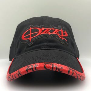 May include: Black baseball cap with a red brim and the word "Ozzy" in red lettering on the front. The brim features the text "Black At The Daddy" in red. The cap is made of a soft material.