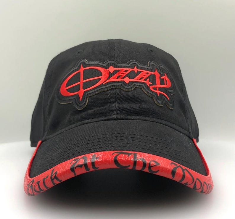 May include: Black baseball cap with a red brim and the word "Ozzy" in red lettering on the front. The brim features the text "Black At The Daddy" in red. The cap is made of a soft material.