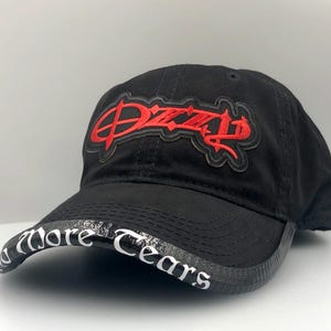 May include: Black baseball cap with the word "Dizzy" embroidered in red on the front. The brim features the words "More Tears" in white lettering. A stylish and casual accessory.