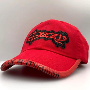 May include: A red baseball cap with the word "Dizzz" in black script outlined in black on the front. The brim has the words "Of A Damon" in black script. The cap is made of a soft material.