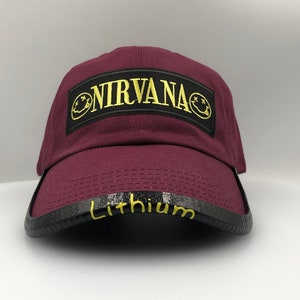 May include: Burgundy baseball cap with a black patch that says "NIRVANA" in gold lettering. The patch has a yellow smiley face on each side. The brim of the cap has a black band with the word "Lithium" written in yellow.