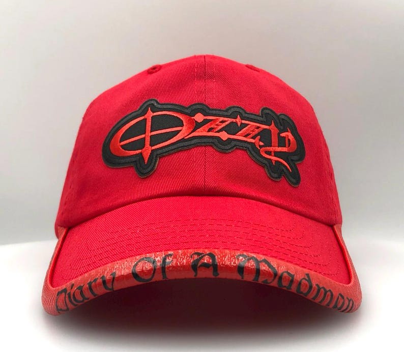 May include: A red baseball cap with the name "Ozzy" in a black and red embroidered design on the front. The brim features the text "Diary of a Madman" in black lettering. The cap is made of a sturdy fabric.
