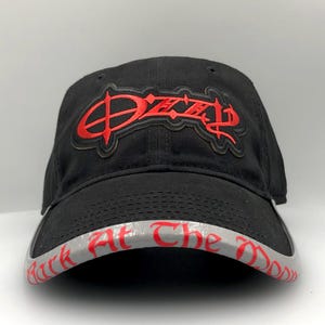 May include: Black baseball cap with the name "Ozzy" embroidered in red on the front. The brim features the text "Bark At The Moon" in red lettering on a gray background. A classic, adjustable hat.