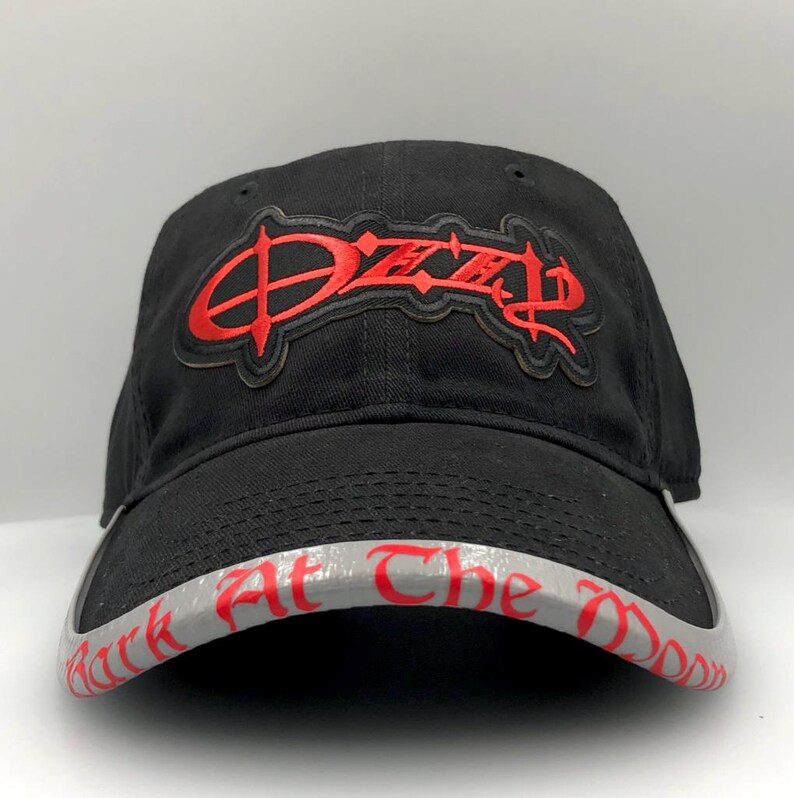 May include: Black baseball cap with the name "Ozzy" embroidered in red on the front. The brim features the text "Bark At The Moon" in red lettering on a gray background. A classic, adjustable hat.