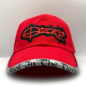 May include: A red baseball cap with the name "Ozzy" embroidered in black and red on the front. The brim is silver with the words "Shot In The Dark" printed on it. The cap is made of a durable fabric.