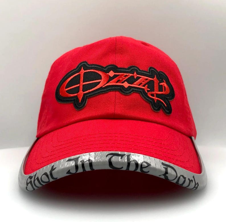 May include: A red baseball cap with the name "Ozzy" embroidered in black and red on the front. The brim is silver with the words "Shot In The Dark" printed on it. The cap is made of a durable fabric.