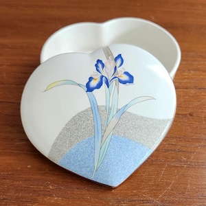 May include: A heart-shaped ceramic trinket box with a white base color. The lid features a blue and white iris flower illustration, accented by a gray speckled curved band and a light blue textured semi-circle at the bottom of the design.