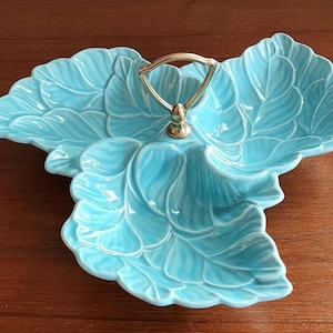 Puede incluir: Una fuente de cerámica turquesa con diseño de hojas. La fuente tiene tres secciones y un asa dorada. El patrón de hojas está detallado con vetas visibles, y la fuente está sobre una superficie de madera.