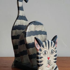 May include: Wooden cat-shaped desk organizer with a black and beige striped pattern. The cat's face has a pink nose and ears, with black stripes and a smiling expression. The organizer has a tall, curved tail.