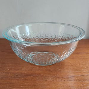 May include: A clear glass mixing bowl featuring a textured exterior pattern of raised circular bubbles of varying sizes. The bowl has a wide, smooth rim and a deep, rounded basin, constructed from transparent glass that reflects light across its surface.