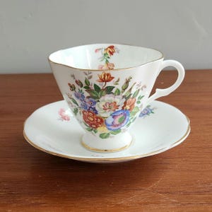 May include: A white porcelain teacup and saucer set featuring a colorful floral bouquet pattern with red, blue, yellow, and green blossoms. The cup has a scalloped rim and a pedestal base, with gold trim detailing on the edges of both pieces.
