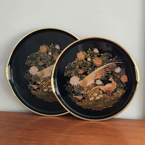 May include: Two round black serving trays with gold rims and light-colored woven handle accents. Each tray features a detailed illustration of long-tailed pheasants among blooming flowers and foliage in shades of gold, pink, and green.