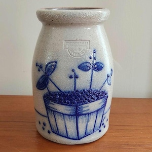 Puede incluir: Un tarro de gres con un acabado azul claro moteado. El tarro presenta una ilustración azul de una cesta llena de bayas y tallos con hojas. El tarro tiene un borde ligeramente ensanchado y un sello rectangular con texto.