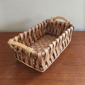 May include: A rectangular woven basket with a light brown color. The basket features a woven base and sides with a handle on each end. The basket is made of natural materials and is suitable for storage or display.