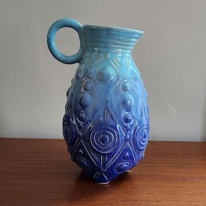 May include: A blue ceramic pitcher with a large handle and a textured surface. The pitcher has a gradient of blue hues, with a lighter blue at the top and a darker blue at the bottom. The design includes raised circles and geometric patterns.