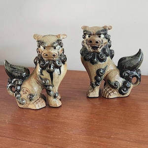 May include: A pair of ceramic Shisa statues, traditional Ryukyu guardian lions. The statues are tan with black and dark green accents, featuring detailed manes and tails. They are posed in a seated position, with open mouths.