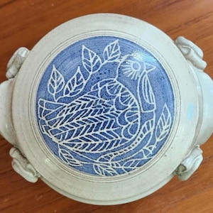 May include: A round, ceramic container with a blue and white bird design. The bird is surrounded by leaves and is centered on the lid. The container has a light beige exterior and small handles on the sides.