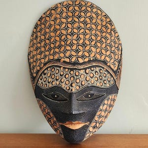 May include: An oval-shaped wooden wall mask featuring intricate pyrography patterns. The forehead displays a repeating floral motif, while the eye area is painted black with white stippling. The lower face is black with a natural wood-toned mouth.