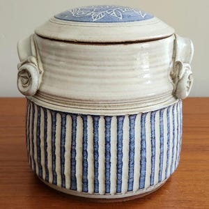 Op de afbeelding: Een keramische pot met deksel. De witte body heeft blauwe verticale strepen, het deksel een blauw bloemmotief. Met decoratieve handvatten, geschikt voor keukengebruik.