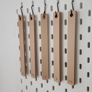 May include: Five beige wooden rulers hanging on hooks on a white pegboard. The rulers are labeled with measurements from 2 to 4.5.