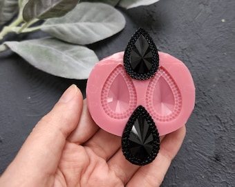 Könnte beinhalten: Rosa Silikonform mit zwei tropfenförmigen Vertiefungen. Zwei schwarze tropfenförmige Strasssteine ​​werden in die Vertiefungen gelegt.