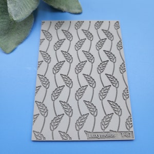 May include: A grey silicone baking mat with a repeating pattern of leaves. The mat has the text "Luxymolds T-87" printed on the bottom right corner.