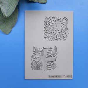 Puede incluir: Un molde de silicona gris con dos diseños. El diseño superior es un sol estilizado con hojas y flores. El diseño inferior es un ciervo estilizado con hojas y flores. El molde está etiquetado como "Luxymolds T-125".
