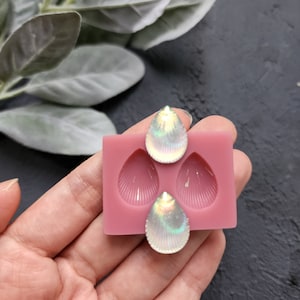 Puede incluir: Un molde de silicona rosa con tres cavidades en forma de lágrima. El molde está diseñado para crear piezas de joyería de resina con un diseño de concha. Dos piezas de resina iridiscente se muestran en el molde.