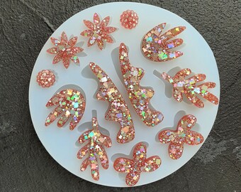 May include: A silicone mold with 10 different shapes, including leaves, flowers, and abstract shapes. The mold is white and the shapes are filled with a glittery pink resin.