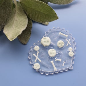 May include: A clear heart-shaped silicone mold with white resin casts of skulls and bones. The mold is on a light blue surface.