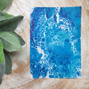 Puede incluir: Impresión artística abstracta con un diseño fluido en tonos azules y blancos, que recuerda a las olas del océano o estructuras celulares. La obra de arte tiene un acabado brillante y se muestra sobre una superficie de madera, con follaje verde en la esquina.