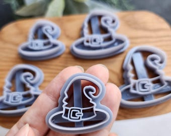 May include: Five gray plastic cookie cutters shaped like witch hats. The cutters are on a wooden surface and a hand is holding one of the cutters.