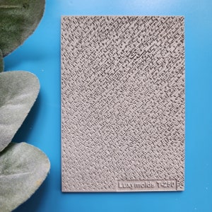 Puede incluir: Un molde rectangular gris claro con una superficie texturizada de pequeños agujeros irregulares. El molde tiene un patrón diagonal y el texto "Luxymolds T-250" está impreso en la parte inferior. El fondo es azul brillante.