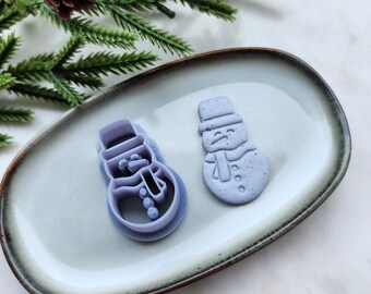 May include: Two purple plastic snowman cookie cutters on a light gray oval plate with a brown rim. The plate is on a white surface.