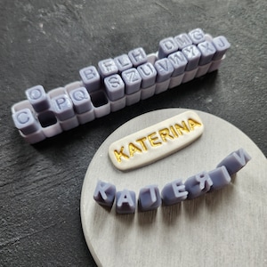 May include: A set of purple alphabet stamps with the name Katerina spelled out in gold letters on a white background.