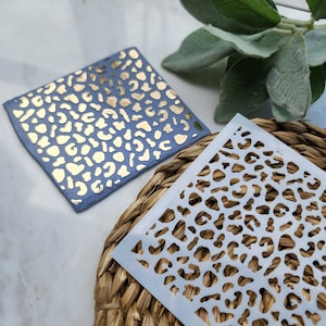 Puede incluir: Dos plantillas de estampado de leopardo, una con pintura dorada y otra con un diseño de corte blanco. Las plantillas están sobre una superficie tejida.