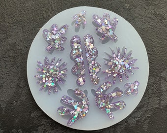 May include: A light blue silicone mold with various shapes, including leaves, stars, and abstract forms. The shapes are filled with iridescent glitter, creating a sparkling effect. The mold is circular and sits on a dark surface.