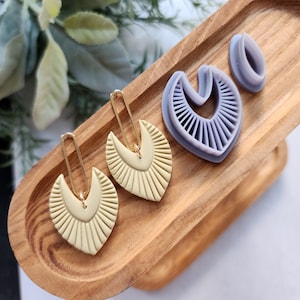 Peut inclure: Trois paires de boucles d'oreilles, deux jaunes et une violette. Les boucles d'oreilles sont en forme d'éventail avec un sommet incurvé. Les boucles d'oreilles sont présentées sur un plateau en bois.