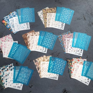 Puede incluir: Una colección de 16 plantillas de arte de uñas azules y blancas con varios diseños geométricos y florales. Las plantillas están dispuestas sobre una superficie gris.