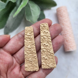 Puede incluir: Un par de pendientes largos y rectangulares hechos de arcilla polimérica beige. Los pendientes tienen un patrón floral detallado en relieve en la superficie.