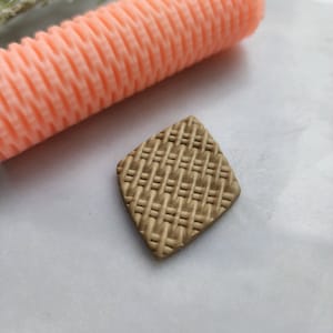 May include: A close-up of a brown diamond-shaped piece of clay with a woven pattern. A pink rolling pin with a textured surface is also in the image.