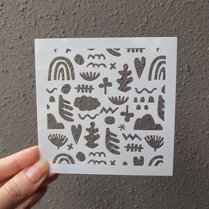 Puede incluir: Plantilla de plástico blanco con un patrón repetitivo de formas abstractas, incluyendo corazones, nubes, arcoíris y hojas. La plantilla está diseñada para manualidades y decoración.