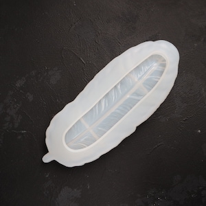 Puede incluir: Molde de silicona blanco con forma de pluma. El molde tiene una superficie lisa y un diseño de pluma detallado.