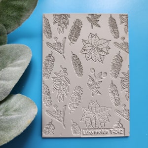 Puede incluir: Un molde para hornear de silicona gris con un diseño en relieve de poinsettias, bayas y hojas. El molde está etiquetado como "Luxymolds T-242".