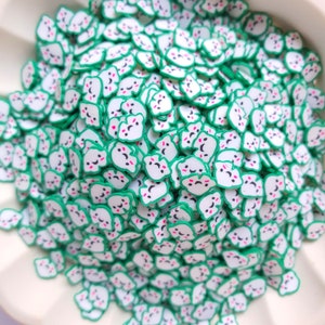 May include: A large pile of white and green resin sprinkles shaped like cartoon faces with closed eyes and a smiling mouth. The sprinkles are perfect for decorating cakes, cupcakes, and other desserts.