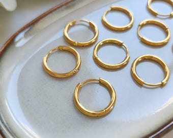 Peut inclure: Plusieurs boucles d'oreilles créoles dorées. Les boucles d'oreilles sont disposées sur un plateau ovale de couleur claire. Les créoles sont de taille moyenne et ont une finition lisse et polie. Les boucles d'oreilles sont de couleur or vif et brillant.