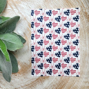 May include: A white fabric sheet with a repeating pattern of red, white and blue hearts. Each heart is decorated with a red, white and blue American flag design.