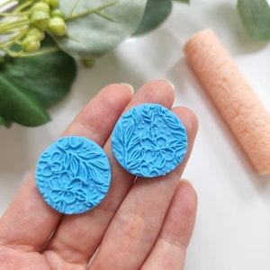 May include: Two round blue polymer clay earrings with a floral design. The earrings are held in a hand.