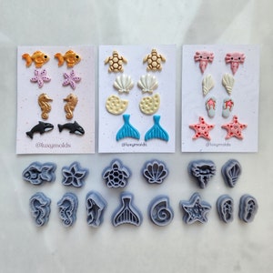 Könnte beinhalten: Ein Set mit 12 Silikonformen zum Herstellen von Polymer Clay Ohrringen. Die Formen zeigen verschiedene Designs zum Thema Meer, darunter einen Seestern, eine Muschel, eine Schildkröte, ein Seepferdchen und einen Meerjungfrauen-Schwanz. Die Formen sind grau und in einem Raster auf einer weißen Oberfläche angeordnet. Der Text "@luxymolds" ist im Bild sichtbar.